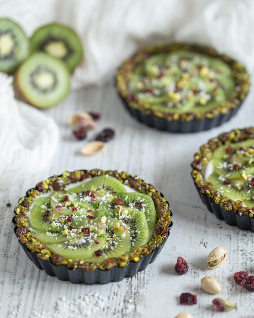 Véganes et sans cuisson, faites le plein de vitamines et d’énergie avec ces superbes tartelettes composées d’une pâte aux fruits secs (pistaches, cranberries, dattes), recouvertes de yaourt végétal et de tranches de kiwi bien fraîches !