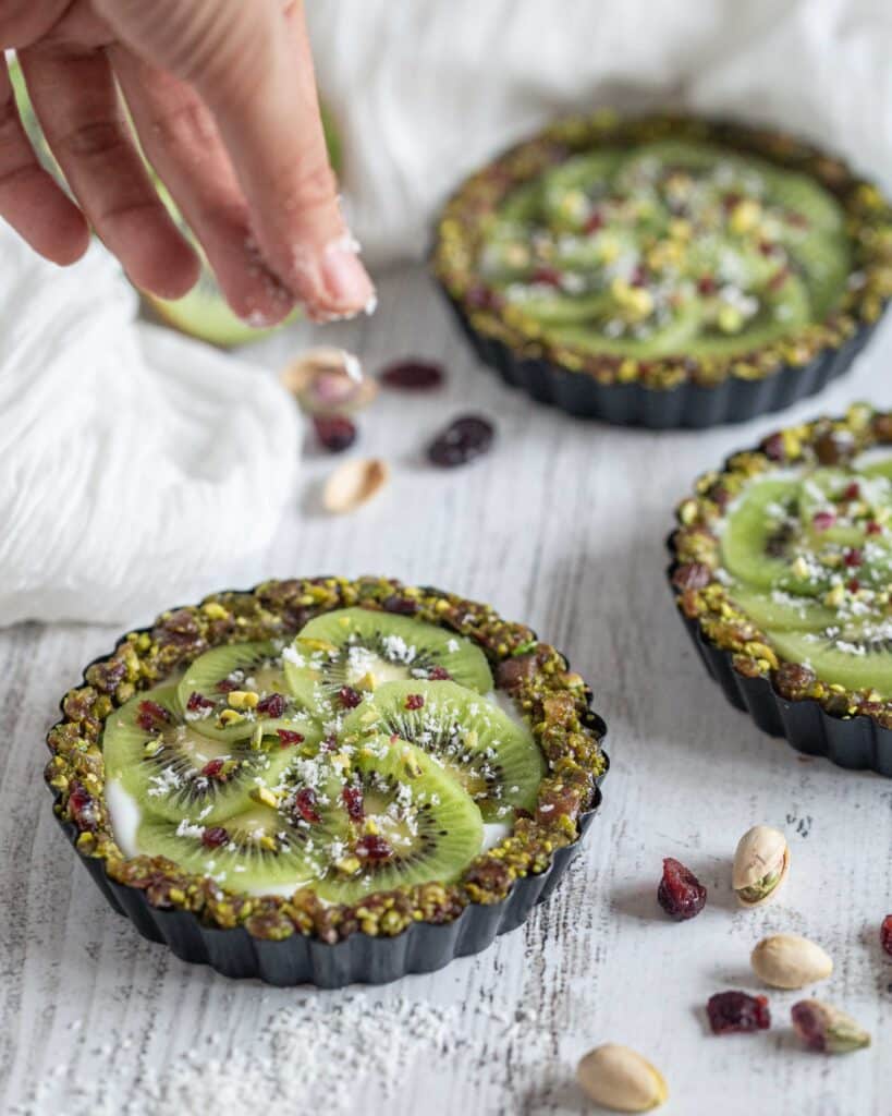 Véganes et sans cuisson, faites le plein de vitamines et d’énergie avec ces superbes tartelettes composées d’une pâte aux fruits secs (pistaches, cranberries, dattes), recouvertes de yaourt végétal et de tranches de kiwi bien fraîches !